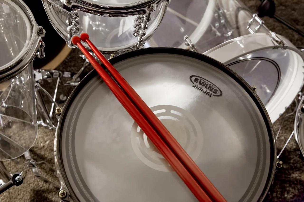 Zwei rote Drumsticks liegen auf dem Fell einer Snaredrum, dahinter die transparenten Kessel des Acryl-Schlagzeugs